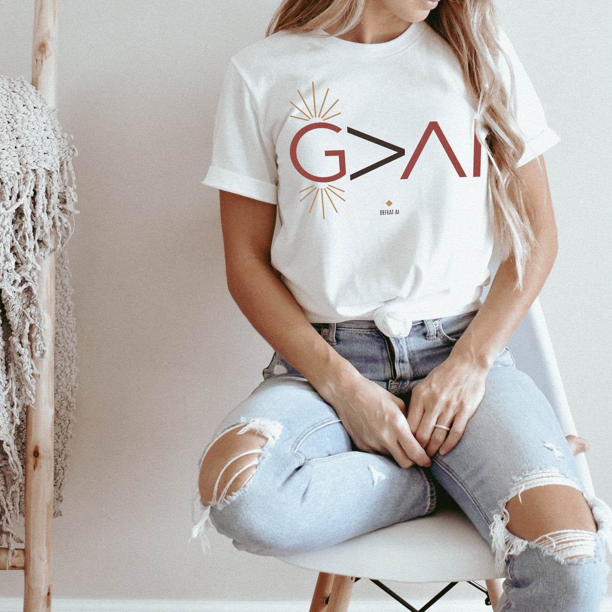 A woman sitting on a white chair wearing a white graphic t-shirt with the text 'G>AI' in red and black with gold sunburst accents, paired with light-wash ripped jeans.