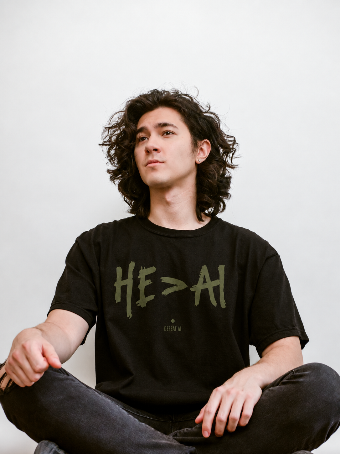 Model seated cross-legged wearing the black HE>AI Christian T-shirt